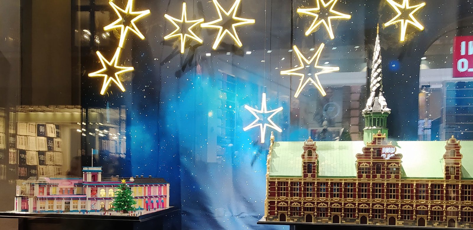 Twee gebouwen van bouwsteentjes in een etalage, met een kerstboompje en verlichte sterren erboven